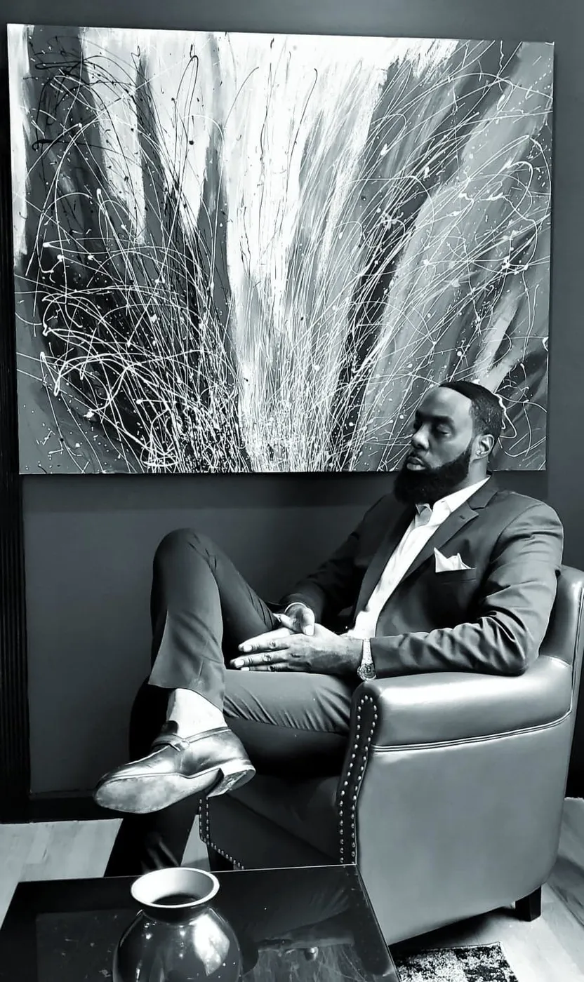Neil Anthony Edwards seated beneath one of his abstract paintings, black and white.